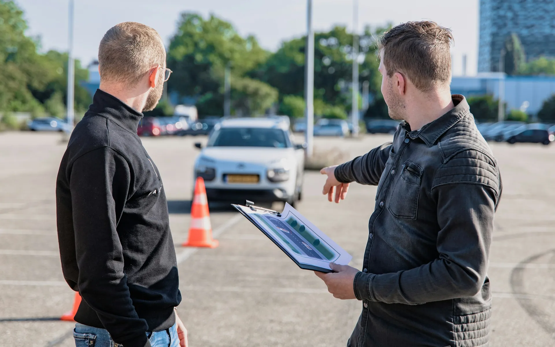 Fahrlehrer erklärt einem Fahrschüler auf dem Übungsplatz eine Aufgabe, im Hintergrund ein Fahrschulauto und Pylonen